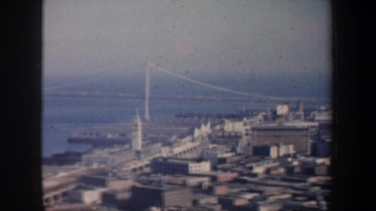 1957 San Francisco home-movie frame showing waterfront buildings, bay water, and a long suspension bridge in soft focus.