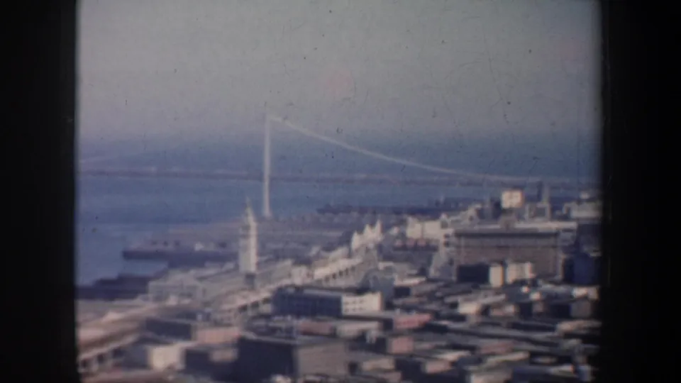 1957 San Francisco home-movie frame showing waterfront buildings, bay water, and a long suspension bridge in soft focus.