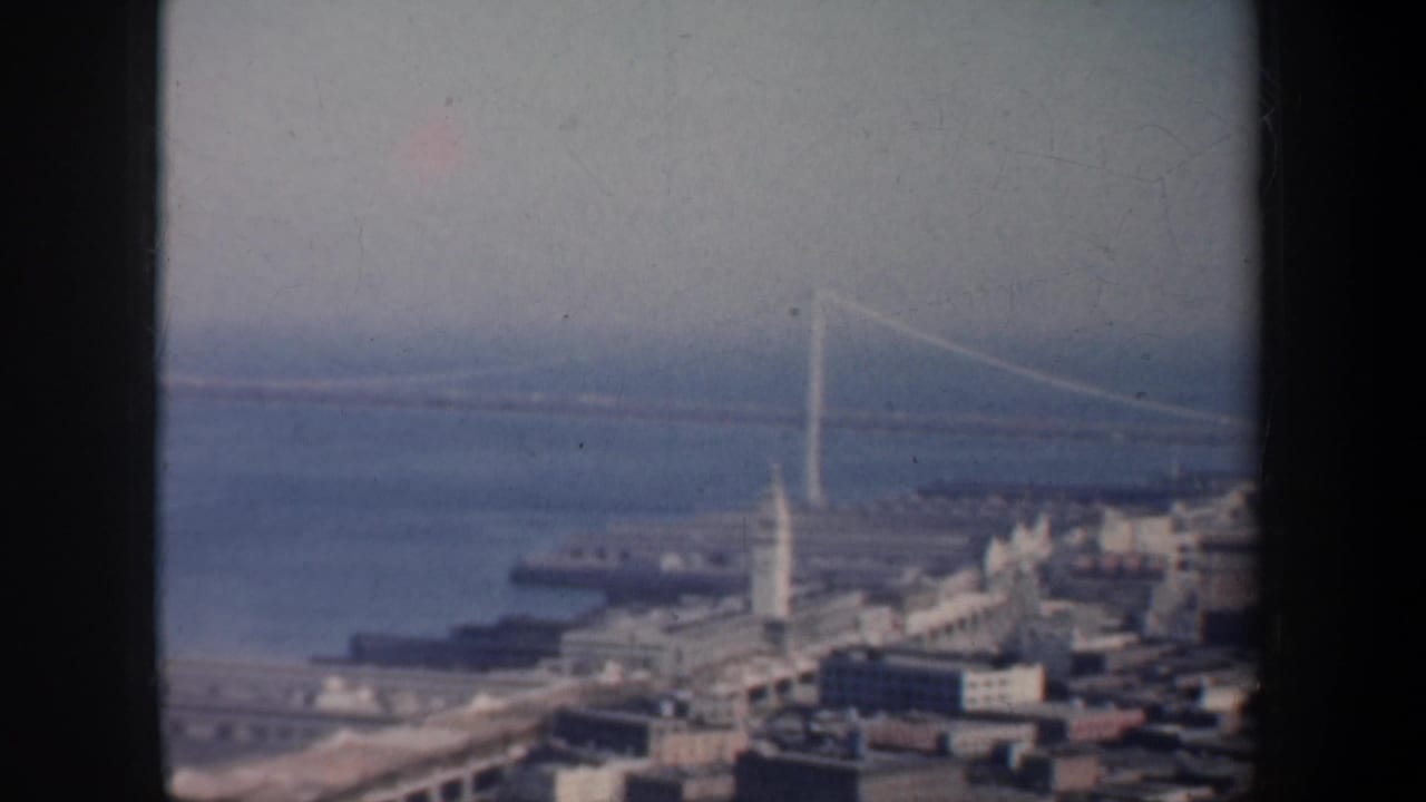 1957 San Francisco frame with a long bridge crossing the bay above piers and clustered waterfront structures.