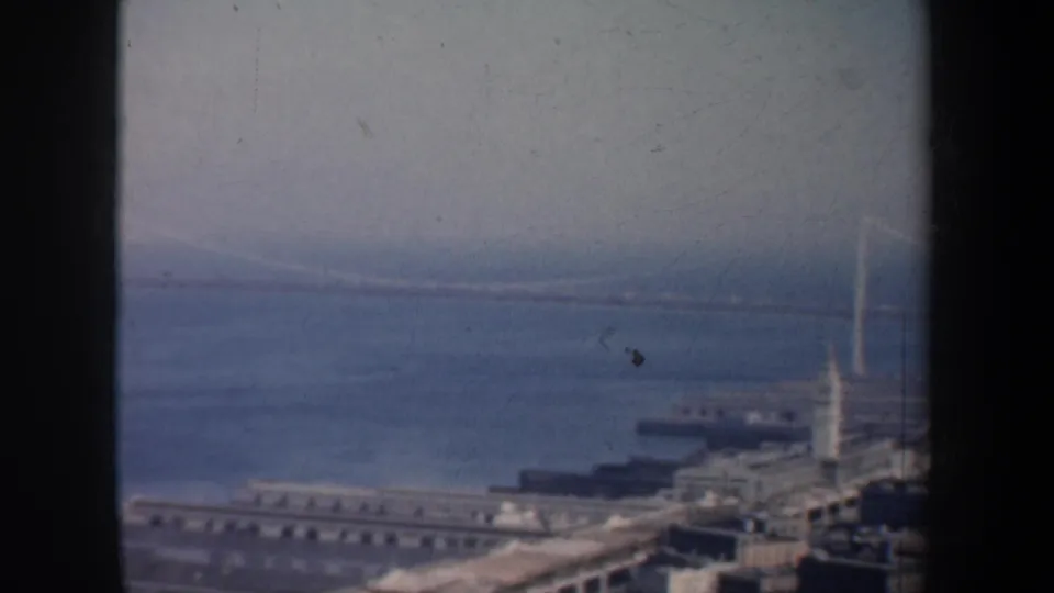 Soft 1957 San Francisco 8mm frame centered on bay water, a bridge tower, and waterfront pier structures.