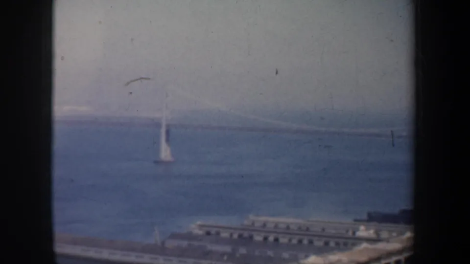 1957 San Francisco bay view with a bridge tower, faint span lines, waterfront pier, and distant shoreline.