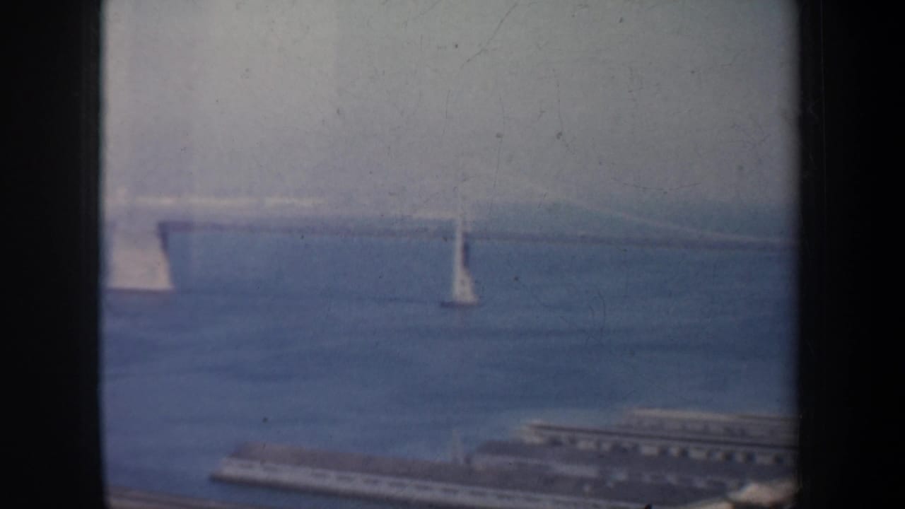 1957 San Francisco Bay view with sailboats on hazy blue water, a distant suspension bridge, and dark vertical framing at the edges.
