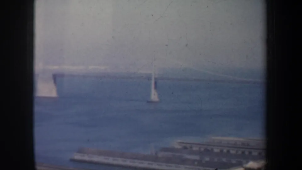 1957 San Francisco Bay view with sailboats on hazy blue water, a distant suspension bridge, and dark vertical framing at the edges.