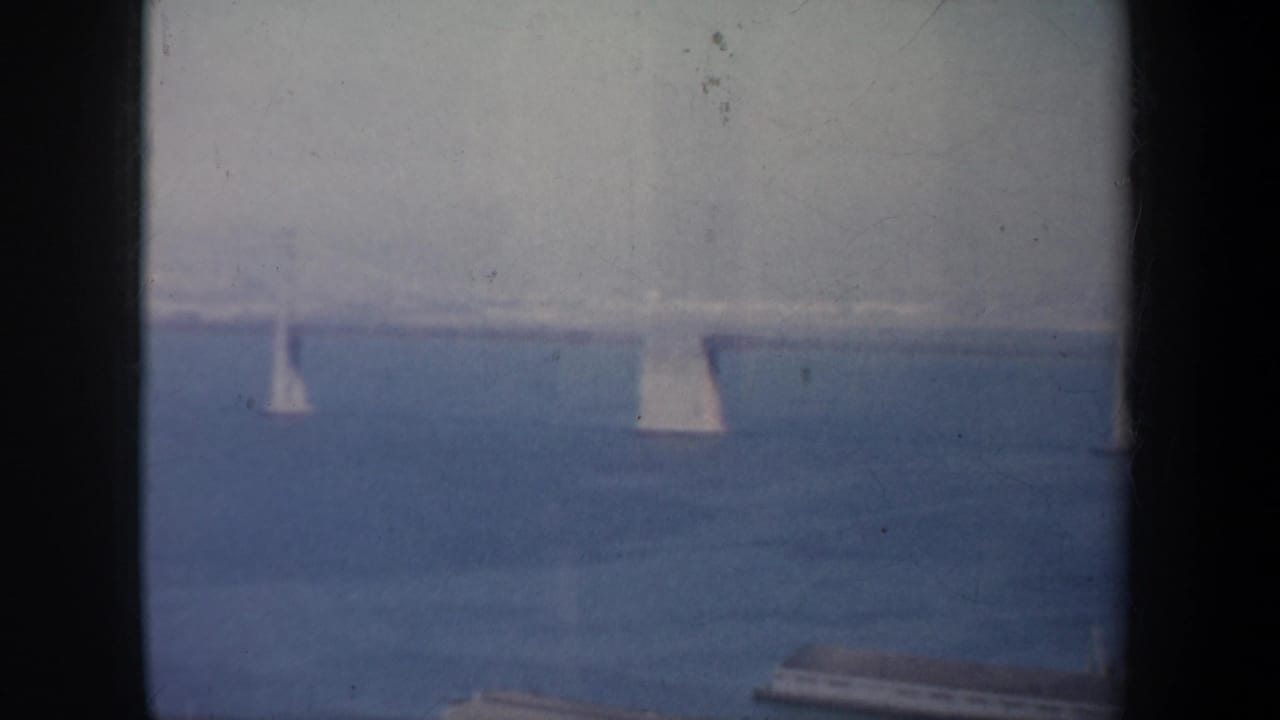 San Francisco Bay sailboats move across blue water beneath a hazy distant suspension bridge in 1957 home-movie footage.
