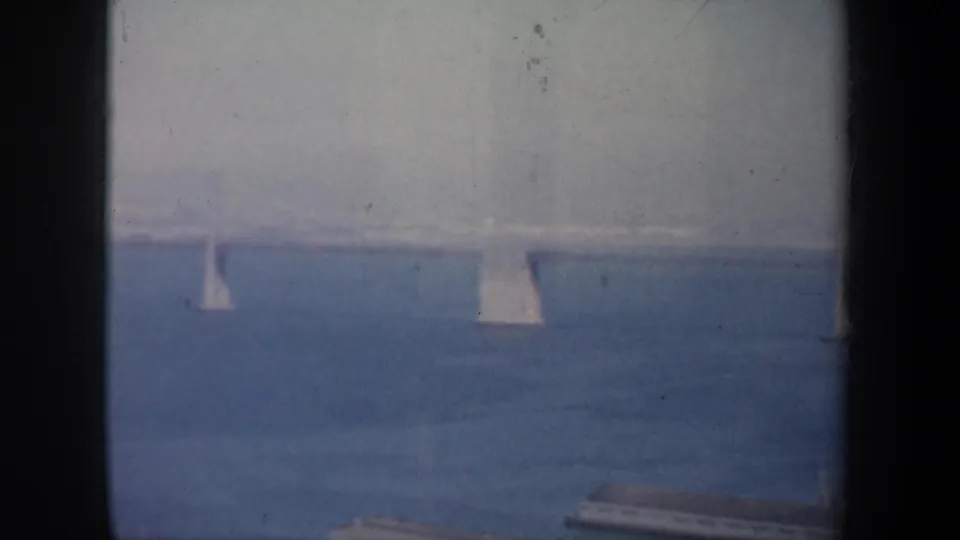 San Francisco Bay sailboats move across blue water beneath a hazy distant suspension bridge in 1957 home-movie footage.