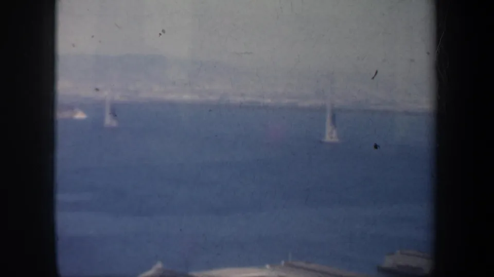 1957 San Francisco Bay home-movie frame with small sailboats, hazy bridge structure, and foreground waterfront edges.