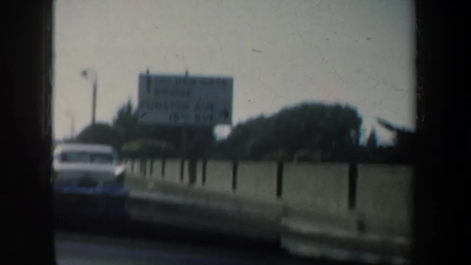 1957 San Francisco roadside footage with a car near a barrier, trees, and a distant sign softened by motion and focus.