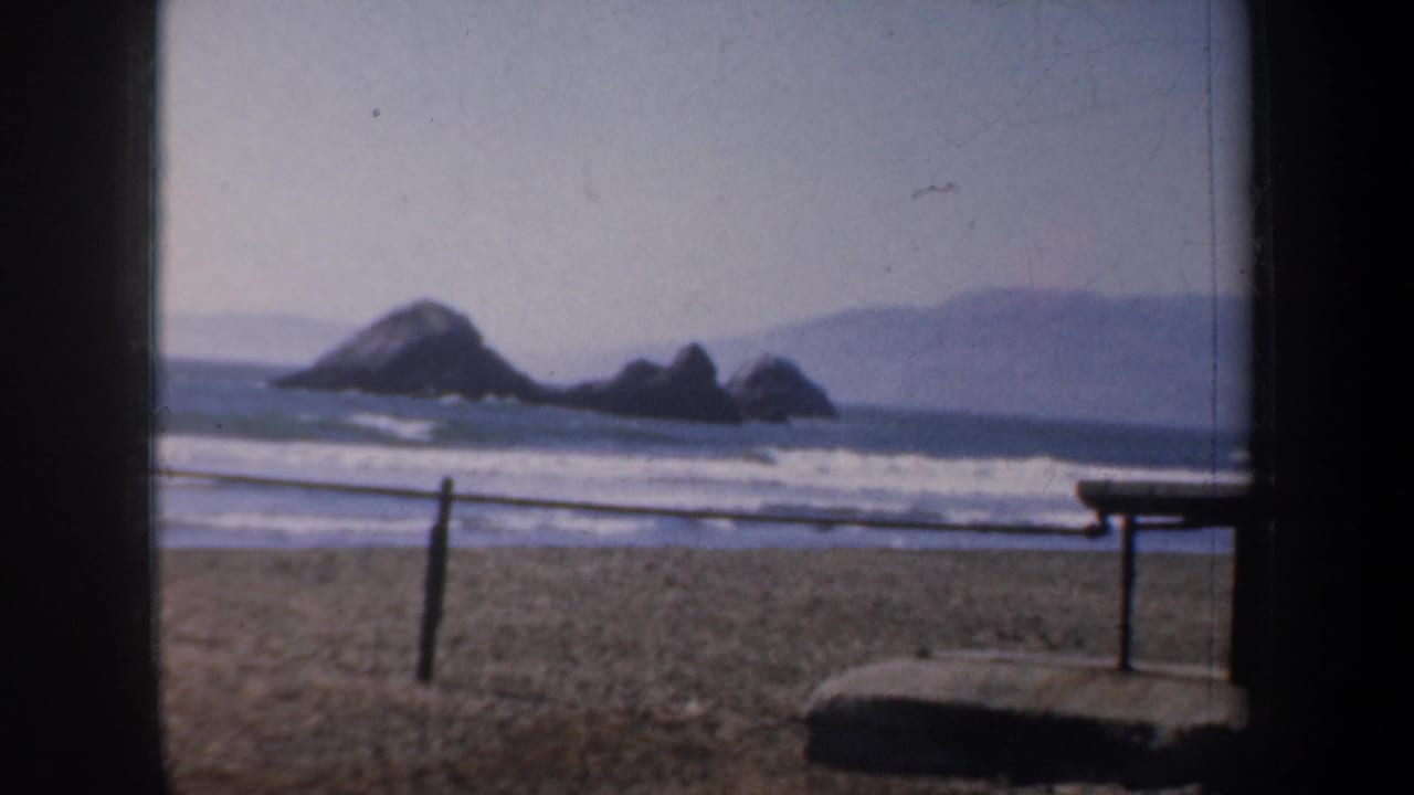 Coastal beach view with offshore rocks, waves, railing, and a bench-like structure.