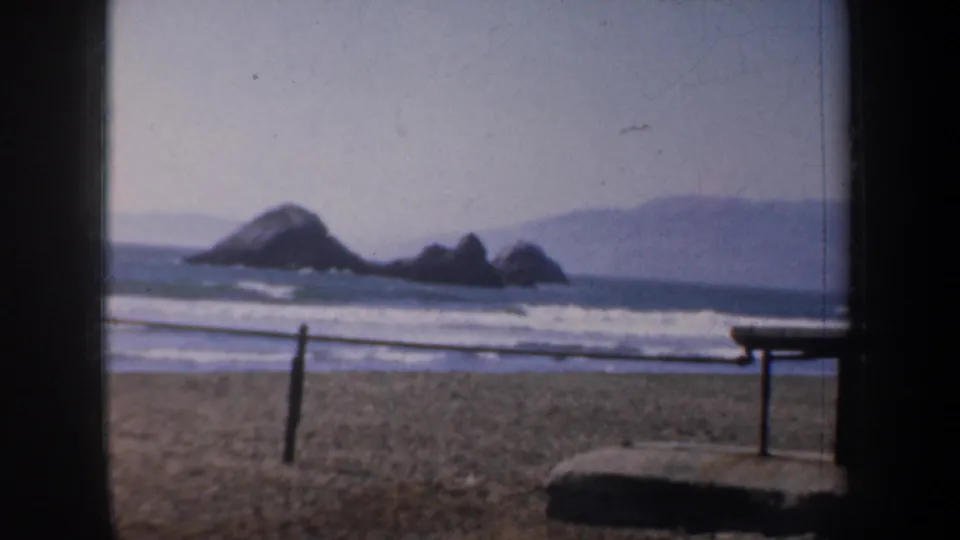 San Francisco coastal view with offshore rocks, rolling surf, beach railing, and distant landforms in soft 8mm color.