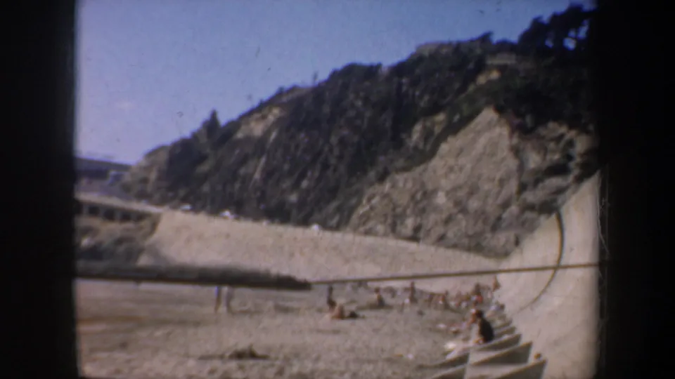 1957 San Francisco beach scene with a rocky cliff, steps, railings, and small groups of people relaxing near the sand.