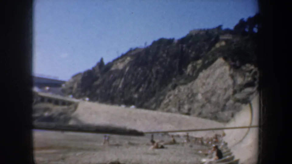 San Francisco coastal beach frame showing a cliff, curved retaining wall, surf, railings, and distant beachgoers in 8mm color.