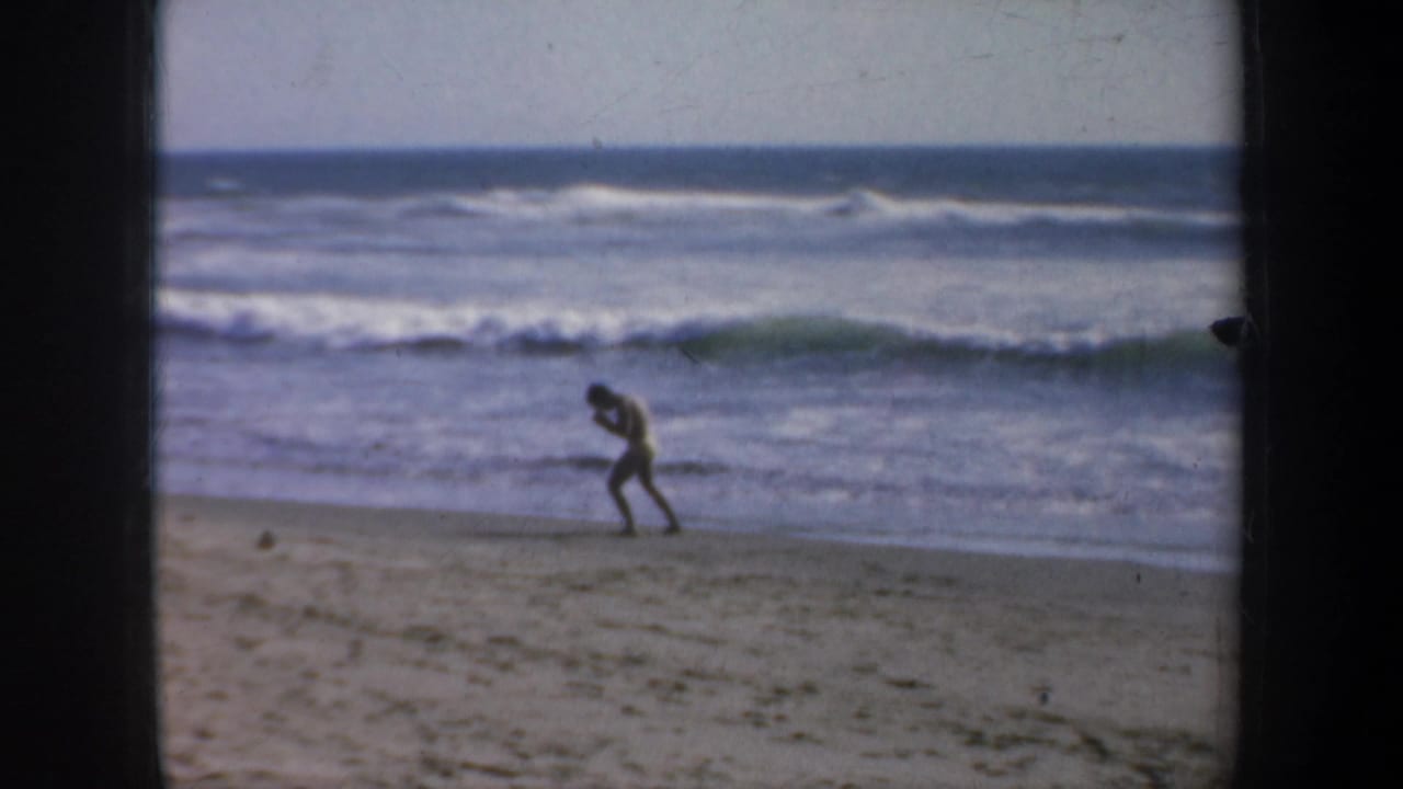 1957 San Francisco 8mm frame showing a man in yellow swimwear shadowboxing near the waterline as surf breaks behind him.
