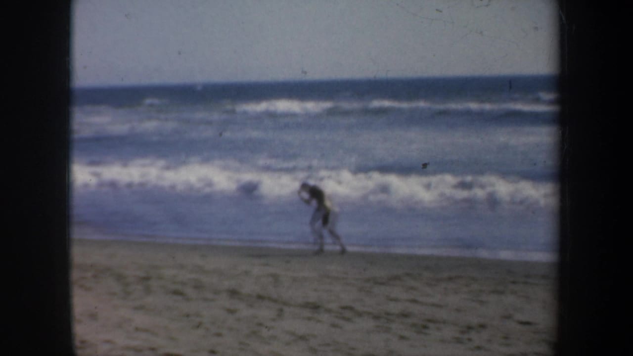 8mm home-movie frame from the supplied 1957 San Francisco beach clip showing the man moving along the sand during boxing practice.