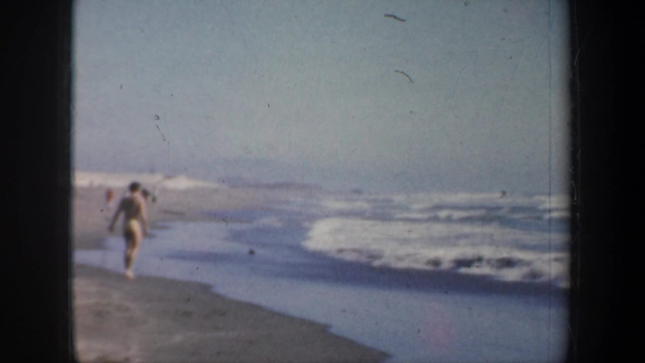 1957 San Francisco beach frame showing the man from behind near the waterline, with pale surf and open sand extending ahead.