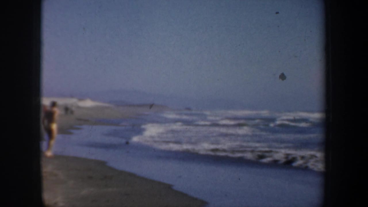 1957 San Francisco home-movie frame showing distant beachgoers near wet sand and surf under a pale sky.