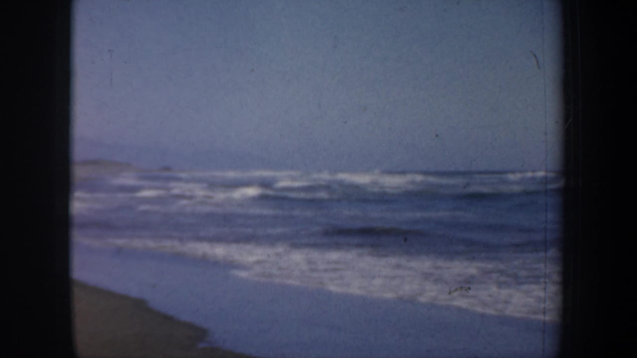 Ocean waves break along a mostly empty shoreline under a pale sky.