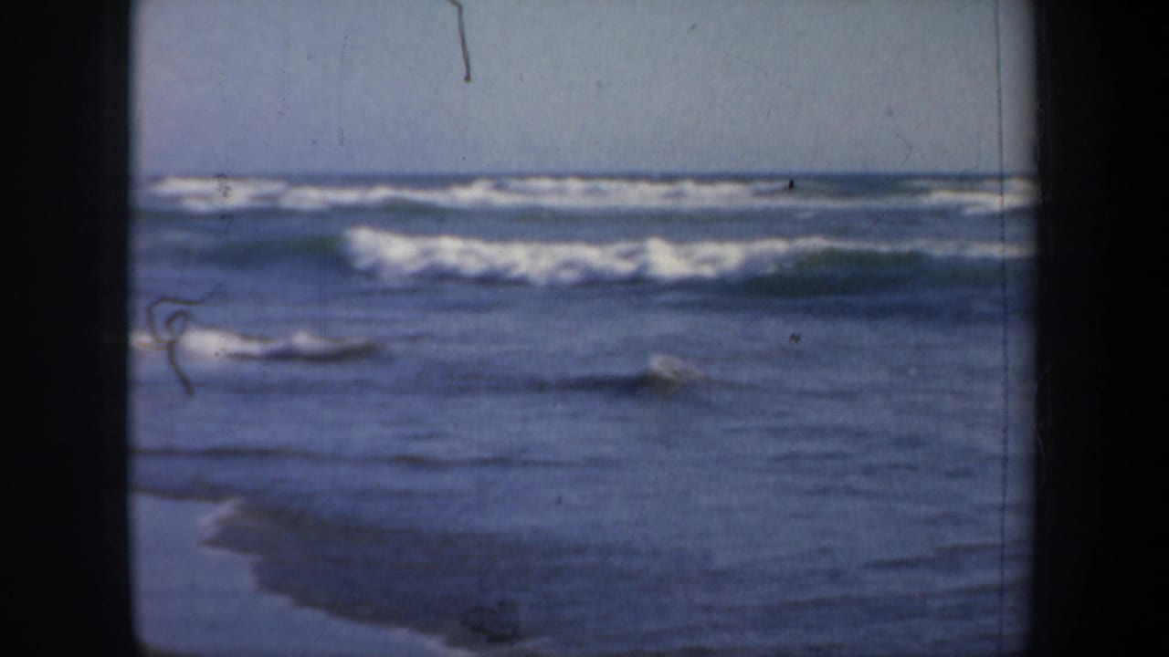1957 San Francisco beach footage frame with low surf, blue water, pale sky, and visible film wear.