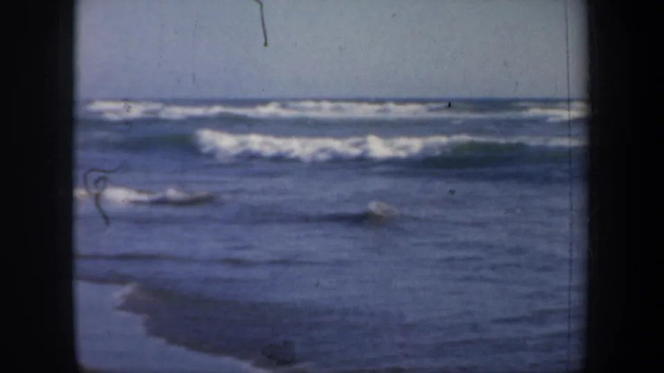 1957 San Francisco beach footage frame with low surf, blue water, pale sky, and visible film wear.