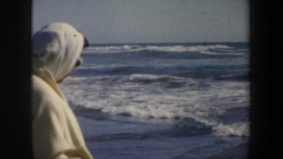 1957 San Francisco beach frame showing a woman in a light headscarf at left, seen from the side or rear as she faces the surf.