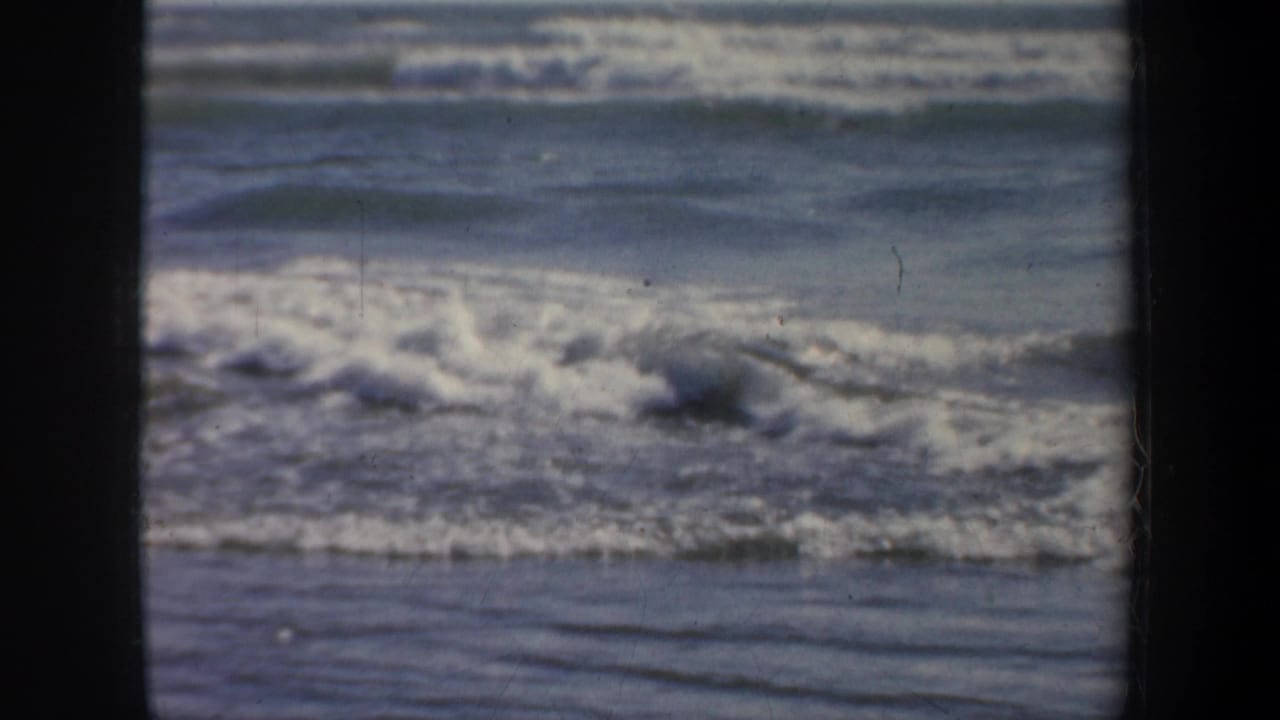 1957 San Francisco 8mm home-movie frame showing white surf and blue-gray ocean under a pale sky with dark film borders.