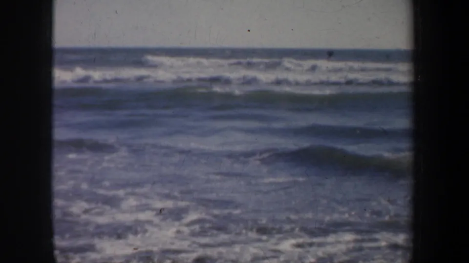 1957 San Francisco coastal home-movie frame with low rolling waves, white foam, muted color, and visible 8mm grain.