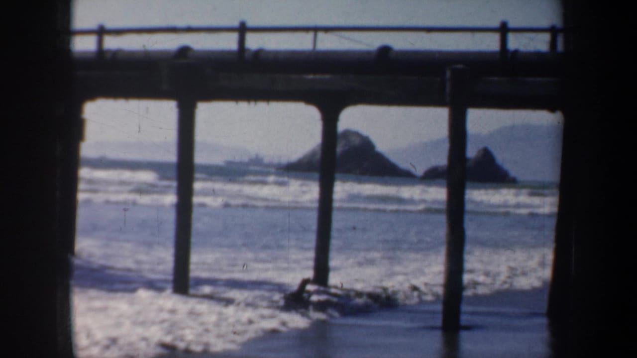 Surf and offshore rocks seen beyond a pier with vertical pilings.