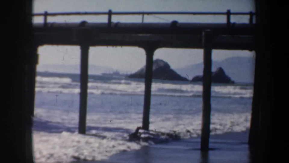 1957 San Francisco coastal view with waves, offshore rocks, distant hills, and a pier structure in soft 8mm focus.