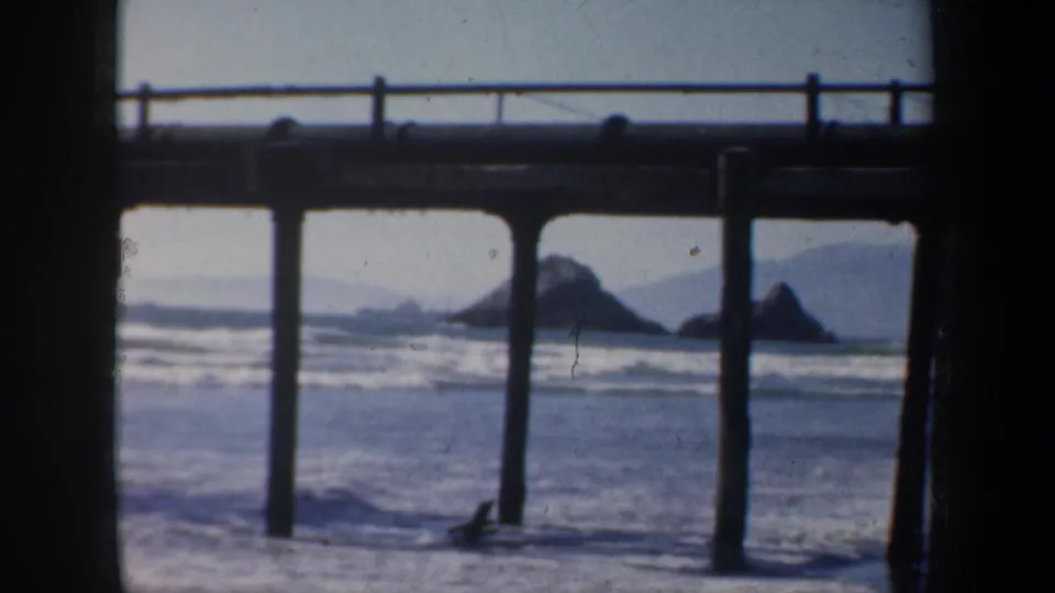 1957 San Francisco 8mm frame of a coastal pier with pilings, pale surf, offshore rocks, and hazy hills beyond.