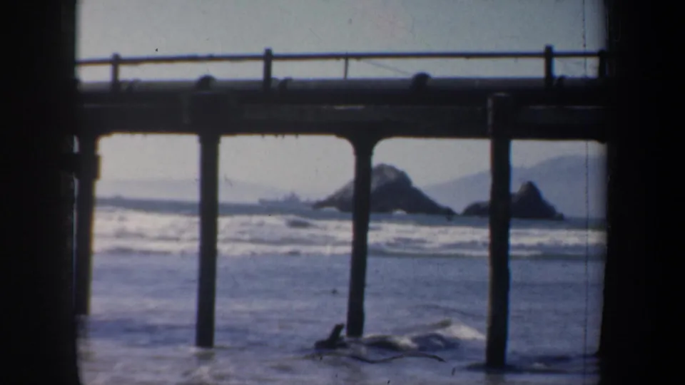 1957 San Francisco coastline shot with silhouetted pier pilings, rolling surf, offshore rocks, and muted distant hills.