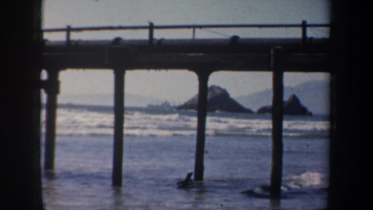1957 San Francisco beach view from beneath a pier, with dark posts, surf, and rocky outcrops in the distance.