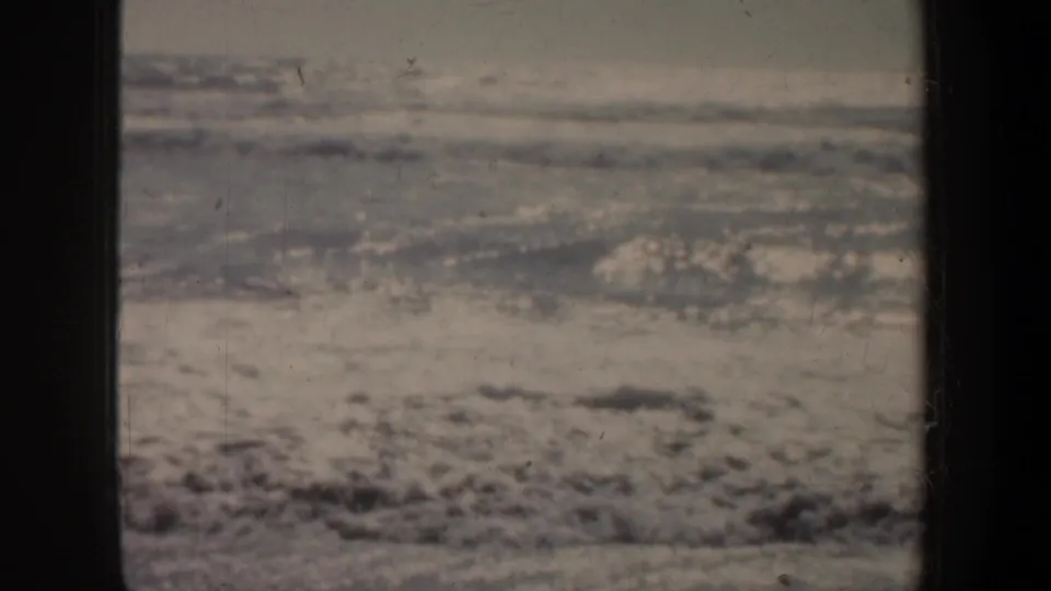 1957 coastal home-movie frame showing pale surf washing across wet sand on a San Francisco beach.