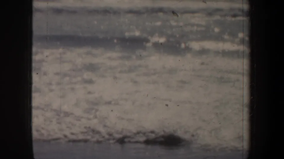 Muted 8mm frame of San Francisco beach waves, with foamy water crossing the foreground and a hazy waterline beyond.