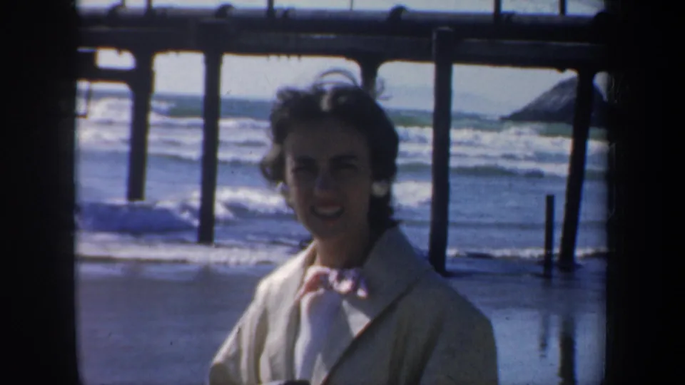 In 1957 San Francisco, a woman faces the camera near wooden pier pilings as ocean waves break behind her.