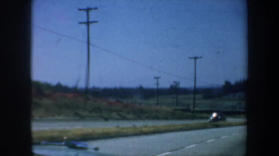 Moving-car view of a San Francisco-area road in 1957, with utility poles, open roadside grass, and a passing vehicle.