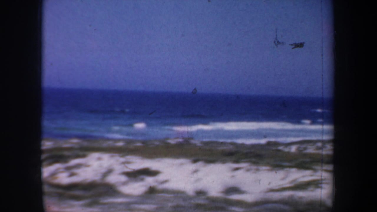1957 San Francisco 8mm beach view with blue ocean, white surf, sandy dunes, and sparse coastal vegetation under a clear sky.