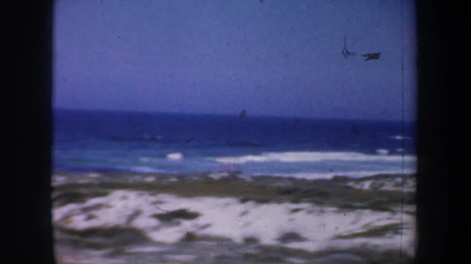 1957 San Francisco 8mm beach view with blue ocean, white surf, sandy dunes, and sparse coastal vegetation under a clear sky.
