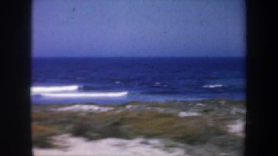 1957 San Francisco 8mm frame with a wide ocean horizon, bands of white surf, pale sand, and coastal scrub in soft focus.
