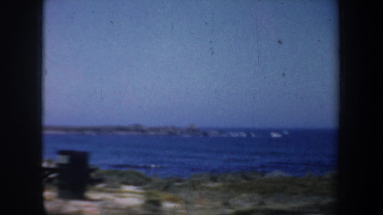 1957 San Francisco coastal home-movie frame with blue water, low beach vegetation, and a dark film border.
