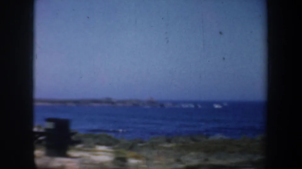 1957 San Francisco coastal home-movie frame with blue water, low beach vegetation, and a dark film border.