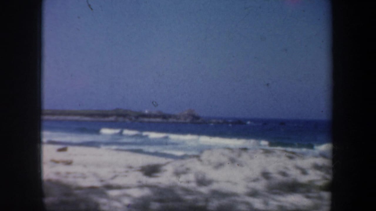 Breaking waves roll onto a pale sandy beach with rocks visible across the water.