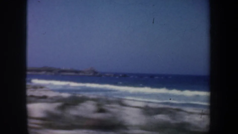 1957 San Francisco coastal frame showing foamy surf, sandy foreground, blue ocean, and distant rocky shoreline.