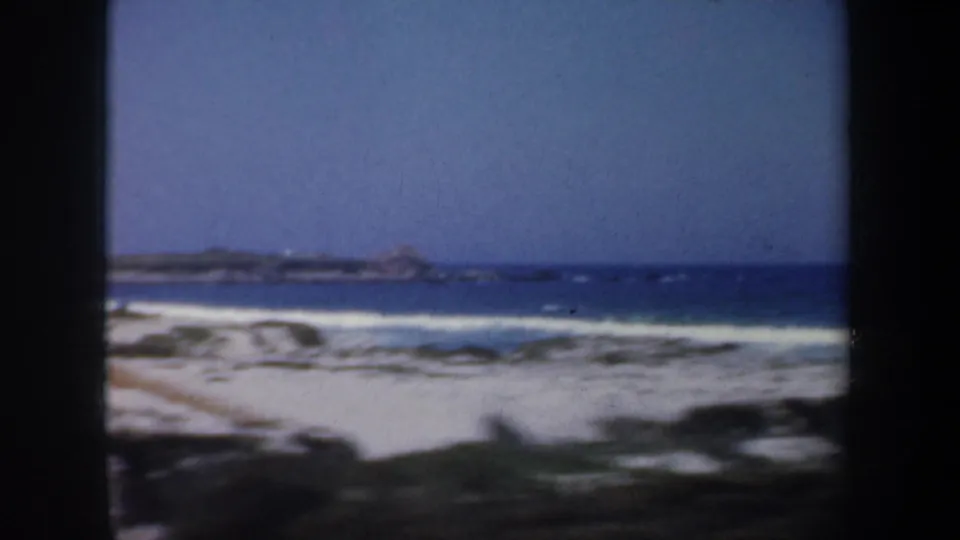 1957 San Francisco beach view with white breaking waves, sand, low vegetation, and a rocky point on the horizon.