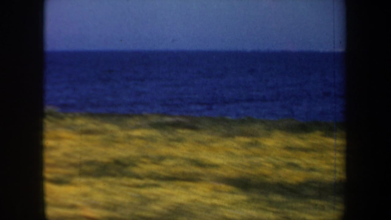 1957 San Francisco-area coastal view from moving 8mm footage, with ocean horizon, pale sky, and grassy shoreline framed by dark vertical edges.