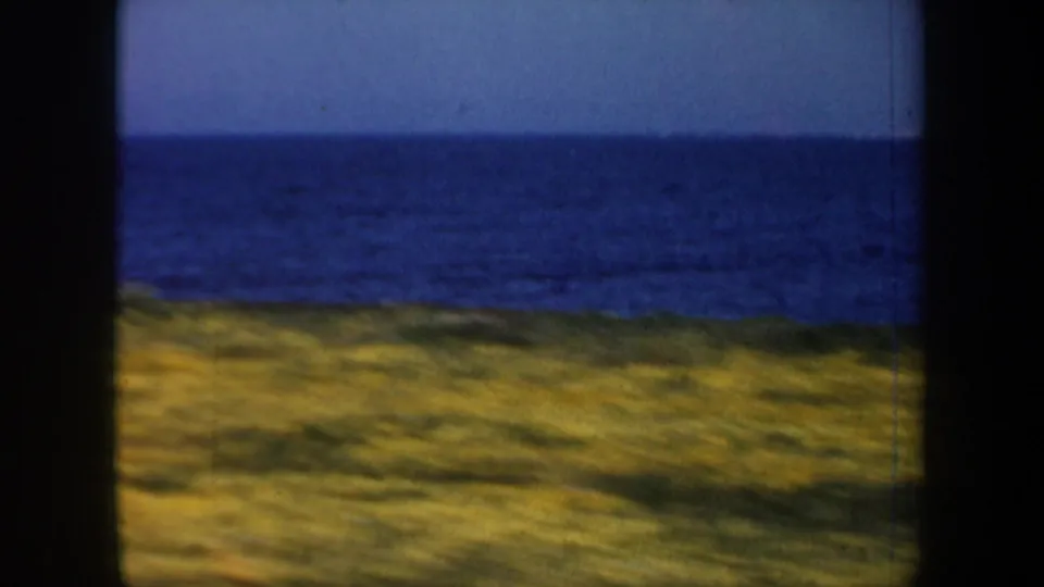 1957 San Francisco-area coastal view from moving 8mm footage, with ocean horizon, pale sky, and grassy shoreline framed by dark vertical edges.