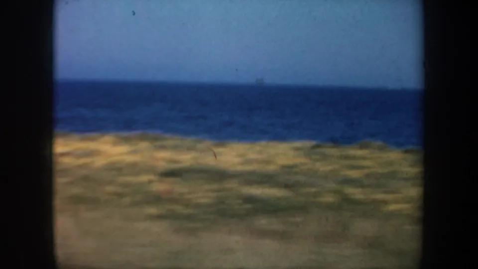 Vintage 8mm frame of the California coast, with blue water, pale sky, and low grassland foreground softened by motion and focus.