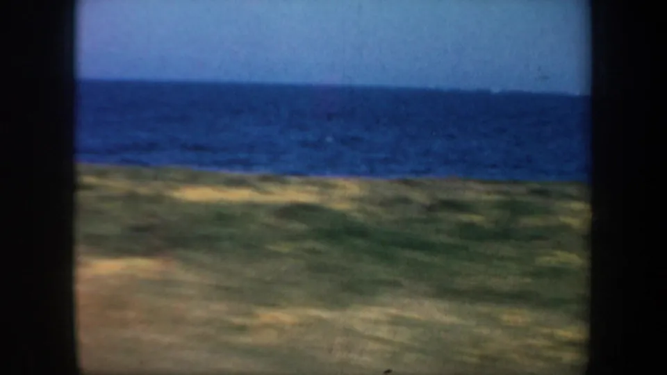 1957 8mm coastal travel frame near San Francisco, showing saturated blue water, pale sky, and blurred grassland from a moving vehicle.