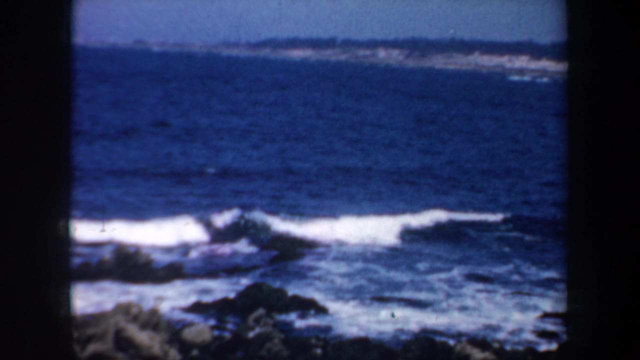 1957 San Francisco coastal home-movie frame showing blue surf breaking against dark rocks, bordered by black film gate edges.