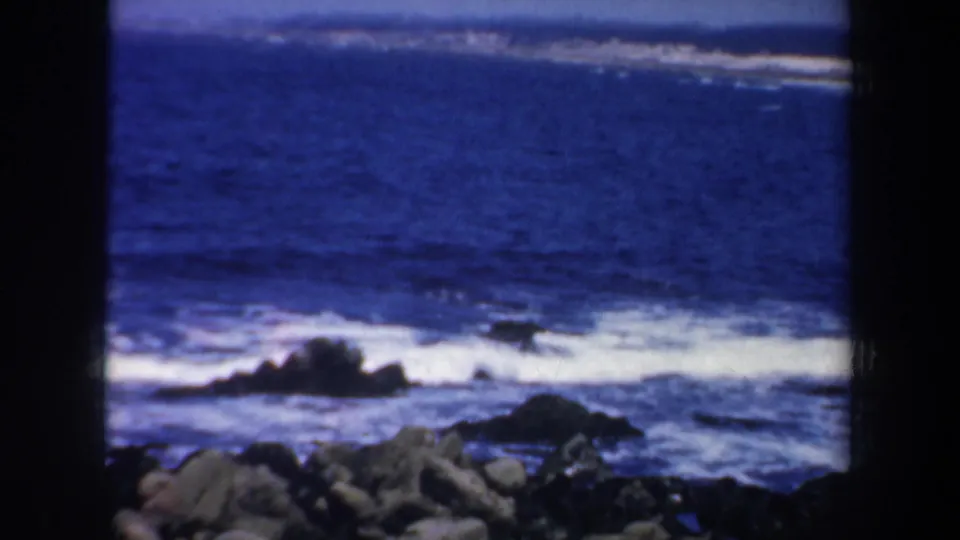 1957 8mm frame of blue Pacific surf and white foam moving around a dark rocky shoreline, with soft focus and film grain.