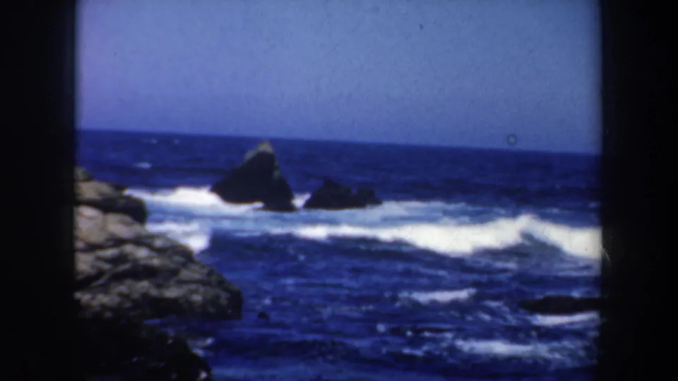 San Francisco coastal home-movie frame showing a prominent offshore rock, blue ocean, white surf, and pale sky in 1957 8mm footage.
