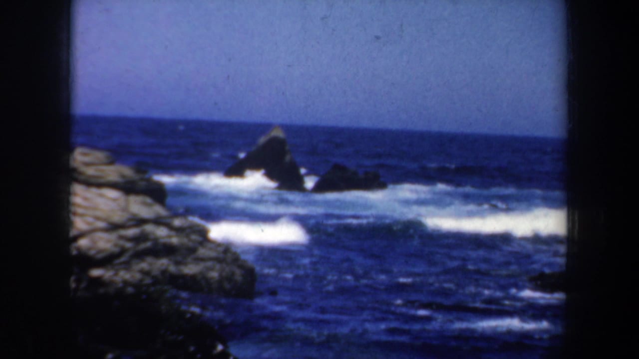 1957 San Francisco 8mm frame showing blue ocean surf breaking around dark offshore rocks and pale coastal stone.